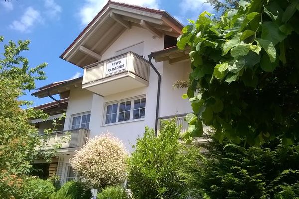 Hinter üppigen Sträuchern steht ein Haus mit einem dreigeschossigen Erker und weißen Sprossenfenstern. Auf dem Balkon im Obergeschoss steht in Großbuchstaben "Fewo Paradies".