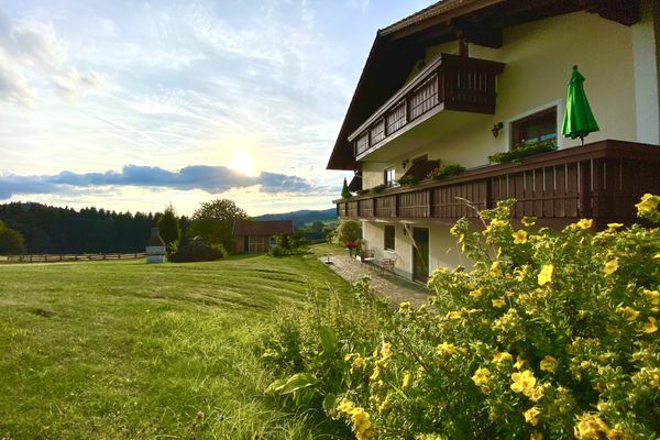 Vor einem dreigeschossigen Haus mit Terrasse und zwei Balkonen steht ein Strauch mit gelben Blüten. Neben dem Gebäude befindet sich eine eingezäunte, gepflegte Wiese mit einem Gartenhaus und einem kapellenartigen Gebäude, dahinter erkennt man die weite Landschaft.