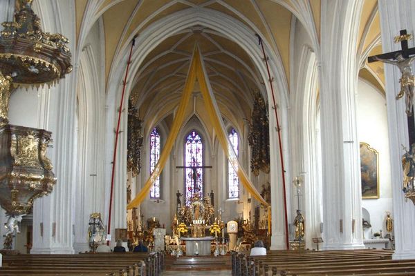 Eindrucksvoller Innenraum der Wallfahrtskirche Bogenberg mit Pfingstkerzen, prunkvoller Kanzel und Bankreihen. Über dem Altarraum hängen gelb-weiße Bahnen von der Netzgewölbedecke