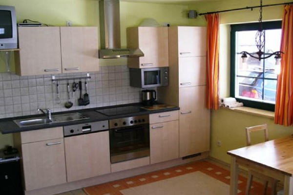 Helle Holz-Küchenzeile mit Spülmaschine, Mikrowelle und Wasserkocher. Neben dem Fenster stehen ein Esstisch und ein Stuhl. Die Vorhänge sind rotgemustert. Auf einem Wandbord steht ein TV-Gerät.
