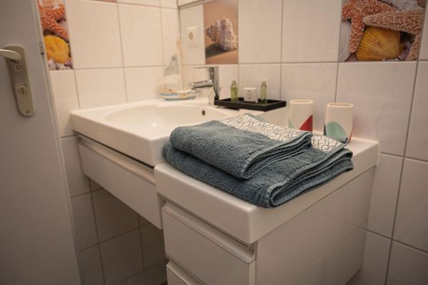 Im Badezimmer stehen ein Waschbecken mit weißem Unter- und Nebenschrank, auf denen Handtücher und Badeartikel verteilt sind. Drei Fliesen mit Muschenmotiven setzen Akzente in den hellen Wandfliesen.
