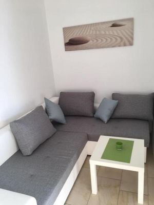 Eine Eckschlafcouch aus weißem Holz mit grauen Polsterauflagen steht vor einem viereckigen weißen Tisch. An der Wand hängt ein längliches Bild mit zwei runden Steinen im Sand.