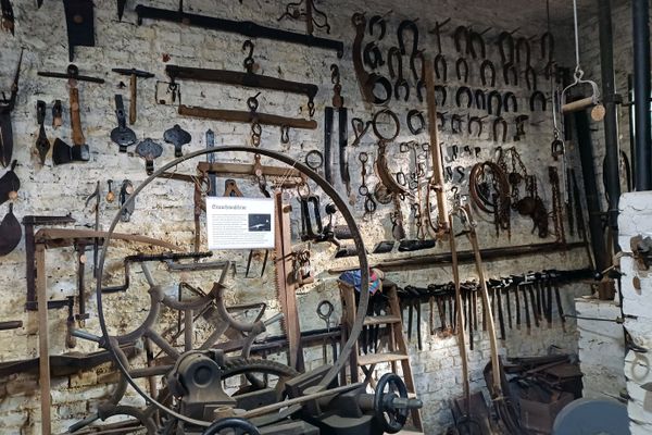 Eine weiß getünchte Steinwand mit unzählig vielen alten Werkzeugen und Hufeisen. Im Vordergrund ist ein eisernes Wagenrad in eine Maschine eingespannt. Auf einem Schild daran steht Stauchmaschine und mehr Text, der nicht zu lesen ist.