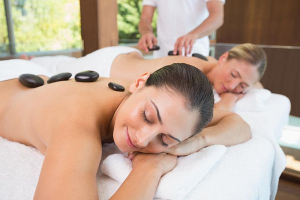 Mit weißen Handtüchern bedeckt liegen zwei Damen im Massageraum auf einer Liege und genießen entspannt und mit geschlossenen Augen eine Warmsteinmassage.