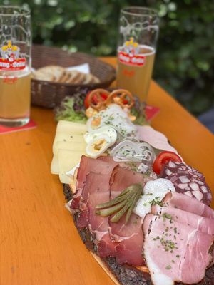 Auf dem Bild ist ein Wurstteller (Brotzeit) auf einem Biertisch zu sehen. Im Hintergrund stehen Biergläser und ein Korb mit Brot.