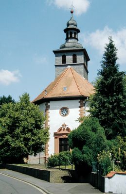 Ein kleines, schlichtes Kirchlein steht zwischen Bäumen auf einer Anhöhe. Eine Treppe führt von der Straße zum Eingangsportal. Der Unterteil mit Kirchenschiff ist weiß verputzt., die Kanten und Fensterlaibungen sind aus rotem Sandstein. Der Kirchturm mit dem achteckigen Haubendachreiter ist dunkelgrau verschiefert. Es ist ein sonniger Tag mit wolkenfreiem Himmel.