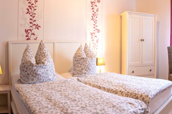 Blick in ein helles Schlafzimmer mit einem weißen Doppelbett, das mit grau-weiß gemusterter Bettwäsche bezogen ist. An der Wand hinter dem Bett sind zwei rot-weiße vertikale Tapetenstreifen mit Blumenmuster zu sehen. Rechts steht ein weißer Kleiderschrank, am Rand rechts sind ein Korbstuhl und ein Tisch angeschnitten. Zwei Nachttischlampen spenden warmes Licht.