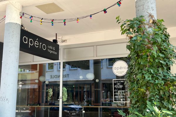 Außenansicht eines Cafés mit schwarzem Schild ‘apéro café & tagesbar‘, einer Säule mit Kletterpflanze und einer Lichterkette mit bunten Glühbirnen