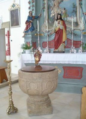 Vor einem Altar mit Christusfigur steht ein steinernes Taufbecken, dessen Deckel von einer Heiligenfigur gekrönt wird.