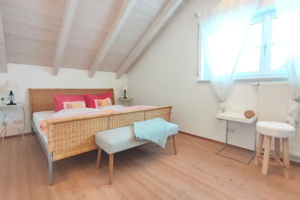 Unter einem offenen, hellen Dachstuhl steht ein Rattan-Doppelbett zwischen zwei Tischchen mit Lampen. Vor dem Bett befindet sich eine Sitzbank und vor dem Doppelfenster steht ein weißes Ablagebord auf einem Edelstahlgestell, daneben ein heller Hocker.