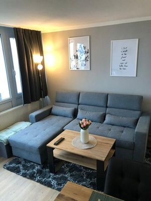 Im Raum befindet sich ein graues Ecksofa, daneben eine Stehlampe. Auf einem schwarz-weiß gemusterten Teppich steht ein Holztisch mit schwarzem Gestell. Am Fenster hängen graue Vorhänge und an der Wand sind in weißem Rahmen eine Zeichnung und ein Spruch zu sehen.