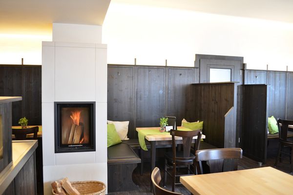 Auf dem Bild sieht man den Kamin in der Gaststube. Auf der rechten Seite, neben dem Kamin, sieht man Tische und Stühle aus dunklem Holz mit grüner Deko. Auch der Eingang ist zu sehen. Auf der rechten Seite ist ein kleiner Teil der Theke zu sehen.