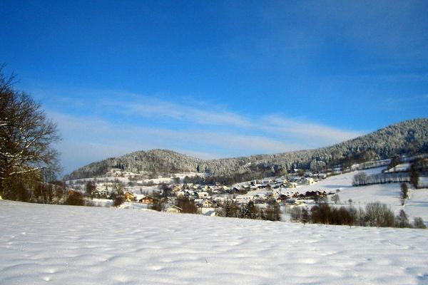 Alternativtext: Eine Ortschaft, eingebettet in schneebedeckte Wiesen, Sträucher und hügelige Wälder.