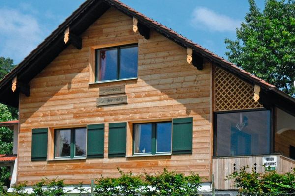 Ein holzvertäfeltes Haus mit grünen Fensterläden trägt den Schriftzug „Landhaus in der Irlau“. Eine große Glasscheibe schützt die Terrasse. Sträucher und Bäume umgeben das Gebäude.