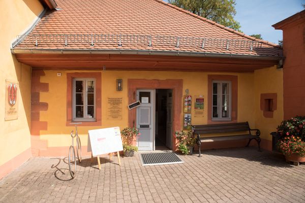 Ein historisches, gelbes Seitengebäude mit roten Ziegeln und Sandsteinumrandungen an Fenster und Tür sowie an Sockel und Kante steht vor dem Betrachter. Der rechte Flügel der grauen Facettentür aus Holz ist geöffnet. Rechts und links der Tür ist jeweils ein Fenster. Rechts vom Eingang hängen an der Wand Dispenser mit verschiedenen Prospekten und darunter steht eine Holzbank. Links des Eingangs steht ein Holzaufsteller mit einem großen Plakat und daneben aus Edelstahl ein lebensgroßes Fahrrad als Fahrradständer. An der Wand des Hauptgebäudes, dass sich links des Seitengebäudes anschließt, hängt das Wappen der Stadt Gedern. Rechts schließt sich ein großer Torbogen an, an dessen Seite ein Blumenkübel steht, genau wie rechts und links des Einganges.