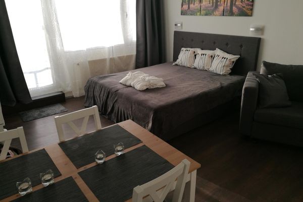 Ein Wohnschlafraum mit grauem Doppelbett und Sofa. Davor steht ein Esstisch, gedeckt mit Tischsets und Teelichtern, umgeben von vier weißen Stühlen.