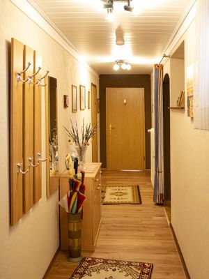 Langer Flur mit Garderobe, Spiegel, Möbeln und Tür am Ende, beleuchtet durch Deckenlampen.
