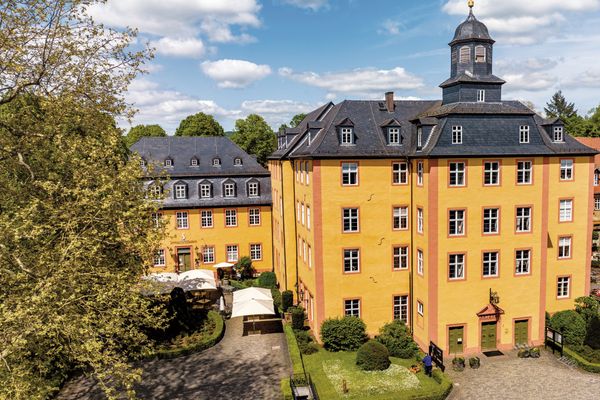 Ein prächtiges, gelbes Schloss mit grauem Schieferdach und markanten roten Fensterleibungen und Hauskanten sowie einem Turm über dem vorgebauten Eingangsbereich ist zu sehen. Gepflegte Grünanlagen drumherum und ein Seitenbau mit dem Schriftzug SchlossHotel Gedern sind vom großen, gepflasterten Hof aus zugänglich.