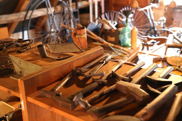 Ein Inneraum des Dorfmuseums zeigt ein Regal auf dem Dachboden. Darauf liegen allerlei Werkzeuge aus Metall, meist Hämmer. Daneben hängt ein kleines, quadratisches Tablett an drei Ketten. Im Hintergrund sind verschwommen Räder mit Speichen zu erkennen und viele Dinge mehr, die unscharf sind.