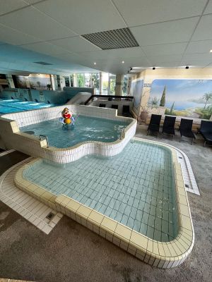Im Kinderbereich des Hallenbades gibt es zwei Kinderschwimmbecken, eines mit einer lustigen Clownfigur, sowie mehrere dunkle Liegen. Die Wände sind mit verschiedenen Motiven und Bildern dekoriert.