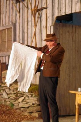 Ein Mann in alter, bäuerlicher Tracht steht lachend da und hält eine knielange, historische Damenunterhose in den Händen, die er neugierig betrachtet.
