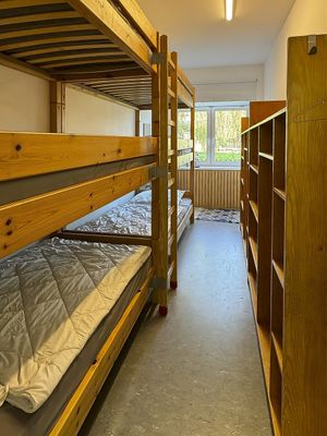 Schmaler Raum mit Holz-Etagenbetten auf der linken Seite und einem Regal auf der rechten Seite. Am Ende des Raums befindet sich ein Fenster.