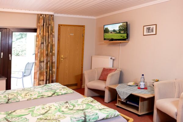 Schlafzimmer mit Doppelbett, zwei Sesseln und kleinem Tisch sowie wandmontiertem Fernseher und Fenster mit Vorhängen.