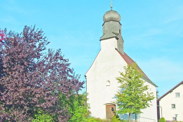 Kleine Kirche mit schindelbedecktem, mehreckigem Kirchturm und Zwiebelkuppel. Links ein blühender Strauch, rechts ein Laubbaum.