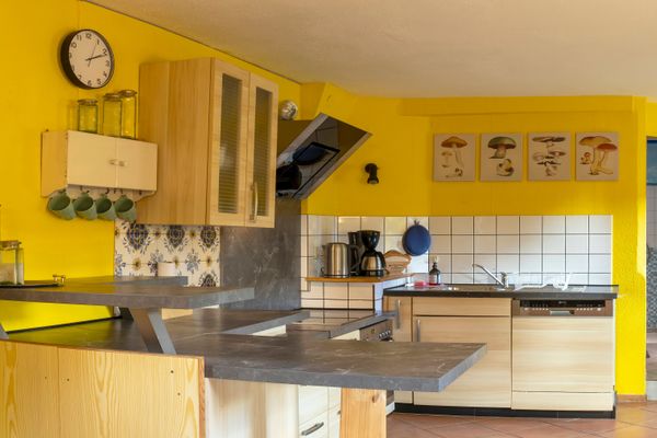 Eine offene Küche mit leuchtend gelben Wänden, terrakottafarbenen Bodenfliesen und hellen Holzschränken mit dunkelgrauen Arbeitsplatten. Ein mehrstufiger Tresen trennt den Arbeitsbereich ab, der mit Spüle, Geschirrspüler, Herd und schwarzer Dunstabzugshaube ausgestattet ist. Im Hintergrund rechts führt ein offener Durchgang in einen weiteren Raum und einen Flur.