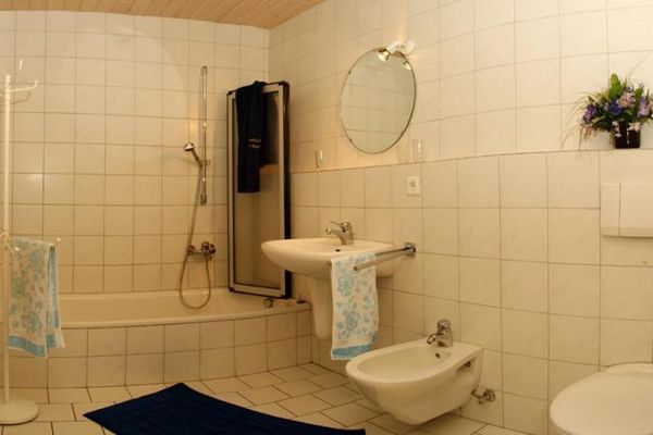 Großes, helles Badezimmer mit Badewanne und Glas-Duschabtrennung. Ausgestattet mit Waschbecken, Bidet, WC und rundem Spiegel. Ein runder, weißer Garderobenständer und ein dunkelblauer Badvorleger ergänzen die Ausstattung.