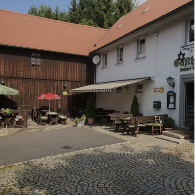 Auf dem Bild ist die Buchwalder Zoigl Stub´n von außen zu sehen. Vor dem Gebäude stehen Bänke und Tische, sowie Sonnenschirme.