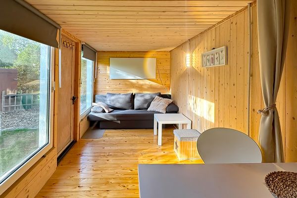 Schmaler Raum mit Holzverkleidung, Sofa am Ende des Raums und kleinem Tisch davor. Links befinden sich große Fenster und eine Tür, rechts ein Tisch mit Stuhl.
