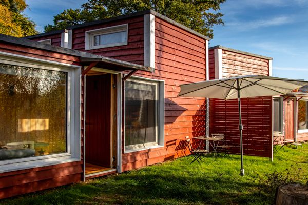 Kleine rote Holzhäuser mit weißen Fensterrahmen und großen Fenstern stehen auf einer Wiese. Davor befinden sich ein Sonnenschirm, ein Tisch und Stühle.