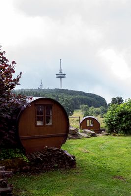 Im Außenbereich auf einer Wiese mit Blick auf den Funkturm auf dem Hoherodskopf, stehen hölzerne Weinfässer. Sie sind eingebettet in eine herrlich grüne Landschaft mit weitem Blick über Wiesen und Waldbereiche.