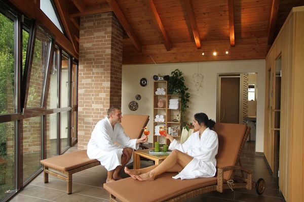 Ein Paar in weißen Bademänteln entspannt im Ruheraum auf Liegen zwischen Fensterfront und Sauna und prostet sich mit Cocktails zu. Im Hintergrund stehen Regale mit Handtüchern und Deko, daneben ist die offene Tür zur Dusche zu sehen.