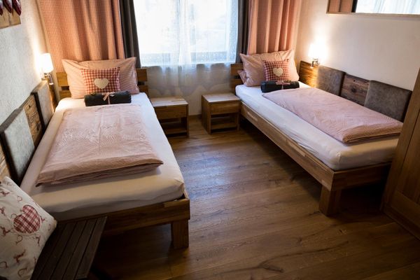 Schlafzimmer in schönem Landhausstil mit zwei Einzelbetten vor einem Fenster mit halbhoher Gardine und rotkarierten Vorhängen.