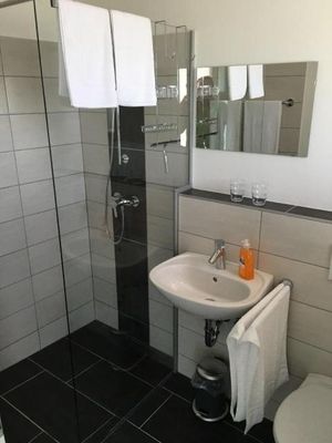 Ein Badezimmer mit einer Dusche mit Glastür, einem Waschbecken, einem Spiegel und einem WC. Die dunklen Bodenfliesen bieten einen schönen Kontrast zu den hellen Wandfliesen.