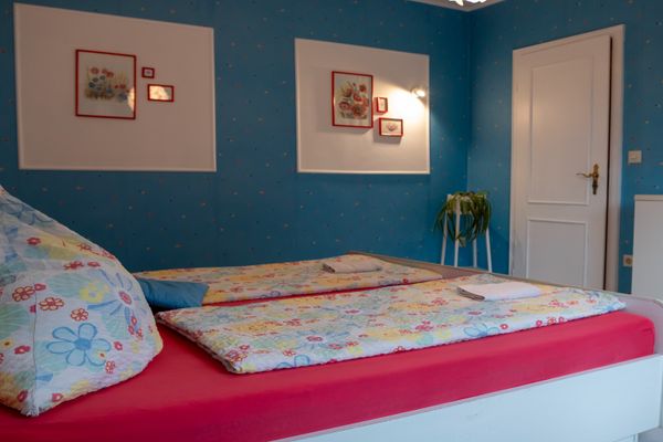 Blick auf ein weißes Doppelbett mit einem leuchtend pinken Spannbettlaken und bunter Bettwäsche mit Blumenmotiven. Auf den Bettdecken liegen gefaltete Handtücher. Die Wände sind blau mit kleinen bunten Punkten und zeigen zwei weiße Felder, in denen kleine Bilder mit roten Rahmen hängen. Rechts ist eine weiße Zimmertür und ein weißer Schrank angeschnitten.