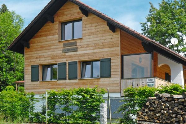 Ein holzvertäfeltes Haus mit grünen Fensterläden trägt den Schriftzug „Landhaus in der Irlau“. Eine große Glasscheibe schützt die Terrasse. Sträucher und Bäume umgeben das Gebäude.
