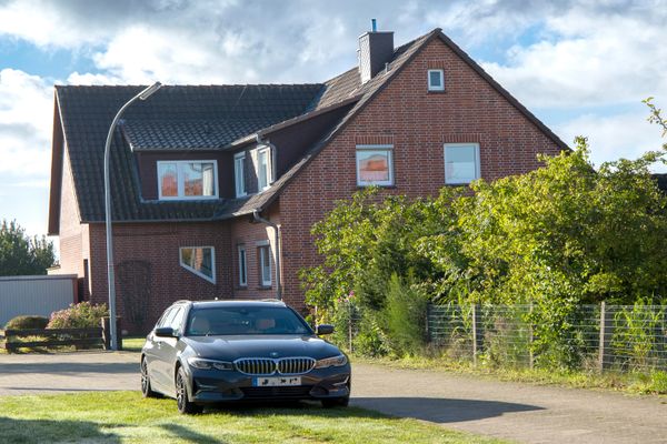 Backsteinhaus mit Satteldach, davor ein geparktes Auto auf einer Zufahrt neben Garten und Zaun.