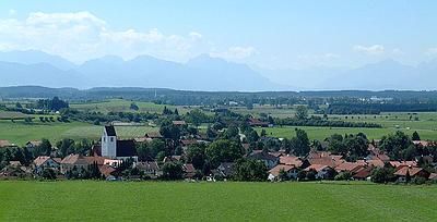 Der Betrachter blickt über ein Dorf hinweg zur Alpenkette am Horizont.