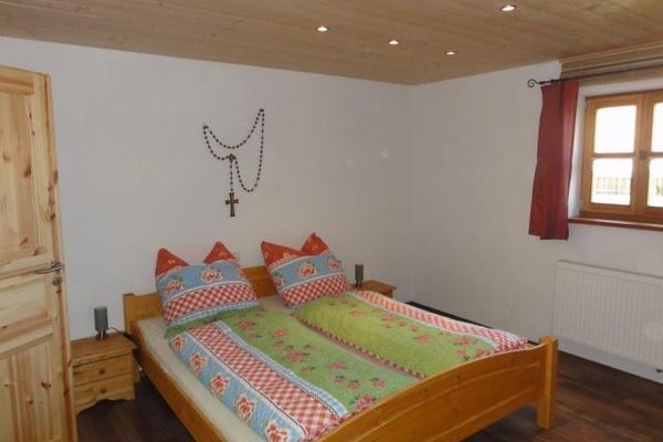 Doppelbett und Nachtkästchen sind aus Kiefernholz. Die Bettwäsche zeigt ein buntes Blumenmuster, und rote Vorhänge hängen am Sprossenfenster. Über dem Bett hängt ein großer Holz-Rosenkranz und Strahler leuchten von der Holzdecke.