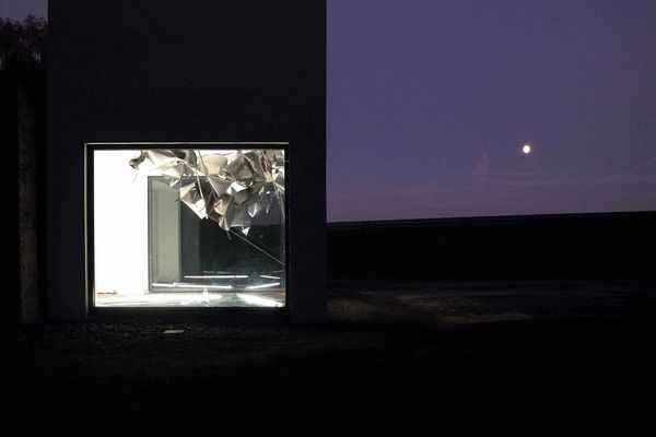 Von außen blickt man bei dunkler Nacht durch das große Fenster eines rechteckigen Turmes. Der gerade so in dunkler Landschaft zu erkennende Turm ist vom Format abgeschnitten, man sieht im Grunde nur das Erdgeschoss und einen Teil des zweiten Stockes. Hinter dem Fenster ist der weiße Raum hell beleuchtet. Ein metallisches Kunstobjekt, das wikt, als wären viele Folien grob gefaltet worden und wie Luftballons an Schnüren schwebend befestigt, ist zu sehen. Der Nachthimmel ist Violett.