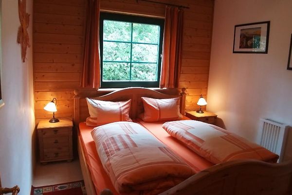 An der Holzwand unter einem Fenster steht ein Doppelbett aus hellem Holz mit zwei Nachttischen und Nachttischlampen. An der Wand hängen zwei versetzt angebrachte Bilder.