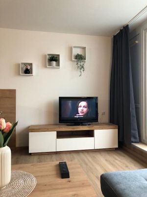 Das TV-Gerät steht auf einem Board in Weiß und Eiche. Darüber hängen drei weiße, viereckige Regale, jedes mit Blumendeko. An der Balkontür sind graue, bodentiefe Vorhänge angebracht. Auf dem Holztisch steht eine Vase mit roten Blumen.