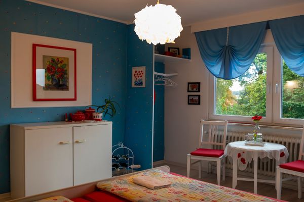 Innenansicht eines Zimmers. Links steht ein weißer Schrank vor einer blauen Wand mit bunten Punkten und einem gerahmten Blumenbild. Rechts befindet sich ein Fenster mit blauen, gerafften Vorhängen, davor eine Sitzgecke aus zwei weißen Stühlen mit roten Polstern und einem kleinen Tisch. Im Vordergrund ist der Fußbereich eines Bettes mit bunter Decke sichtbar. An der Decke hängt eine kugelförmige, weiße Lampe.