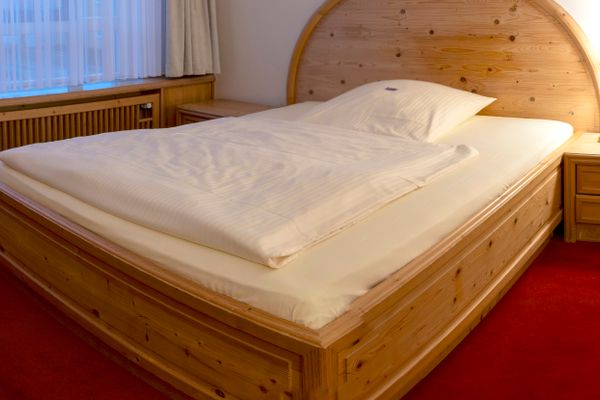 Das Bild zeigt ein Schlafzimmer mit einem großen Holzbett in der Mitte, helle Bettwäsche, einem Nachttisch mit Lampe, großen Fenstern mit weißen Vorhängen und einem roten Teppich.