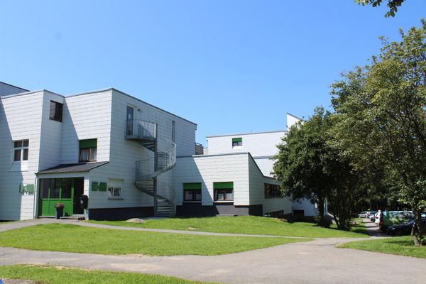Außenansicht einer Appartementanlage mit einer Edelstahl-Außenwendeltreppe. Die Eingangstür und die Rollos sind grün. Vor dem Gebäude gibt es gepflegte Rasenflächen, geteerte Wege und Laubbäume.