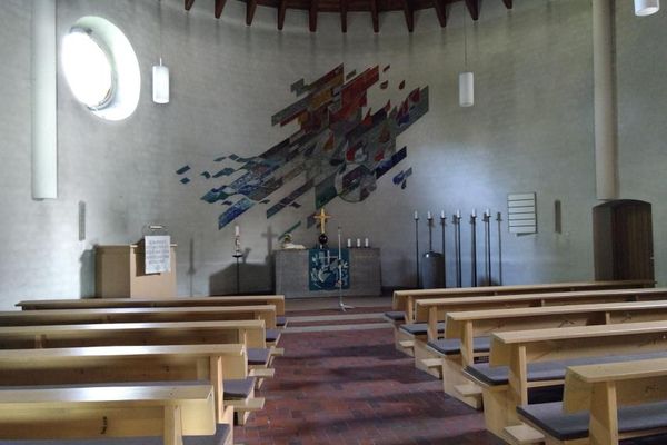 Den steinernen Altar schmückt ein Teppich, ein goldenes Kreuz und weiße Kerzen. An der Wand ist eine Mosaik Grafik in gedeckten Farben angebracht worden.