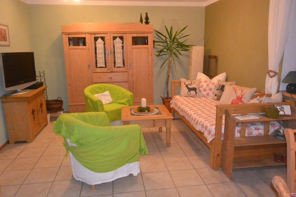 Wohnzimmer mit Fliesenboden, Sofa und Möbeln aus Holz sowie zwei Sesseln die bedeckt sind mit weißen Hussen und grünem Stoffüberwurf.